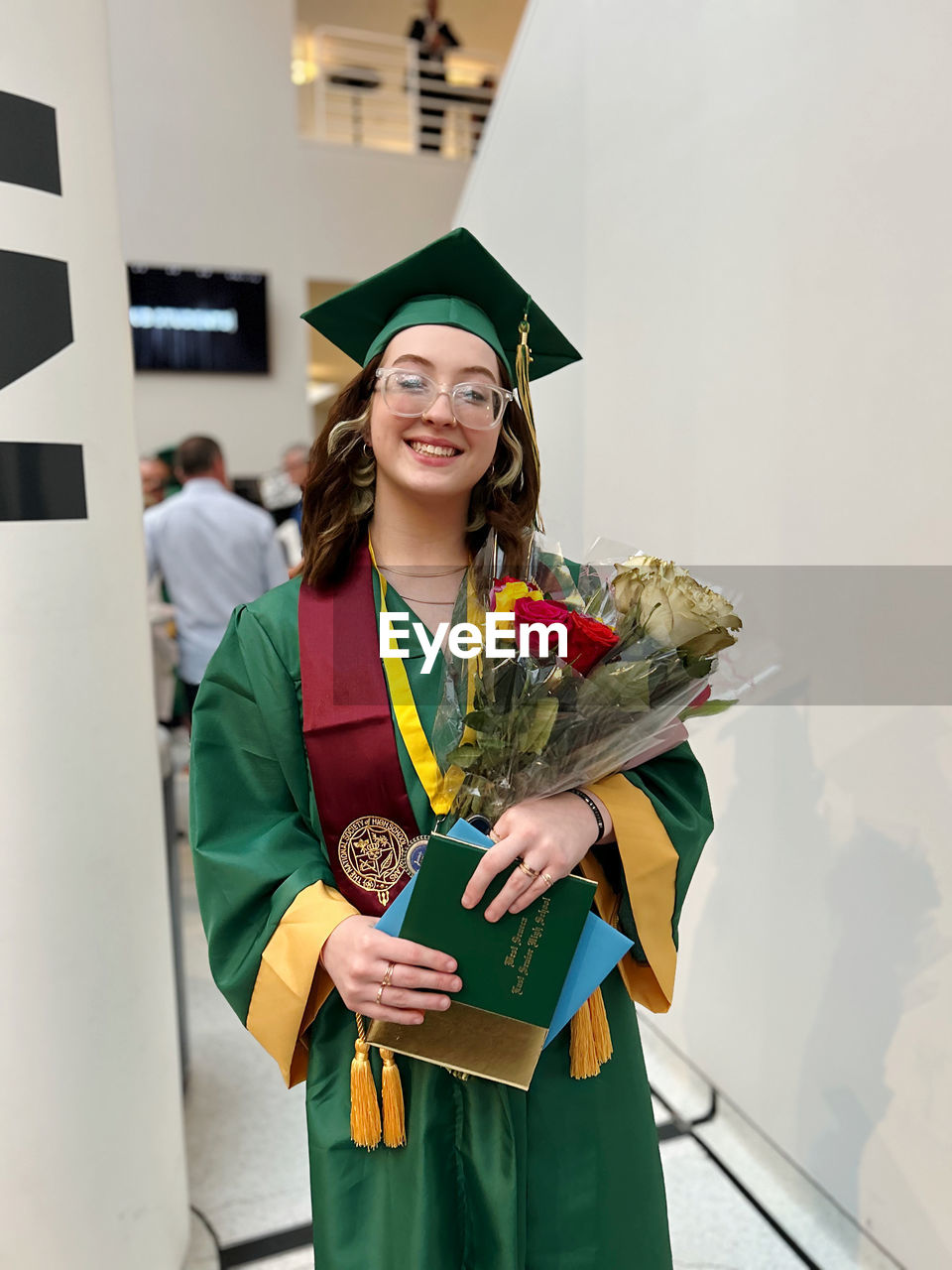Portrait of young woman wearing graduation gown standing against wall