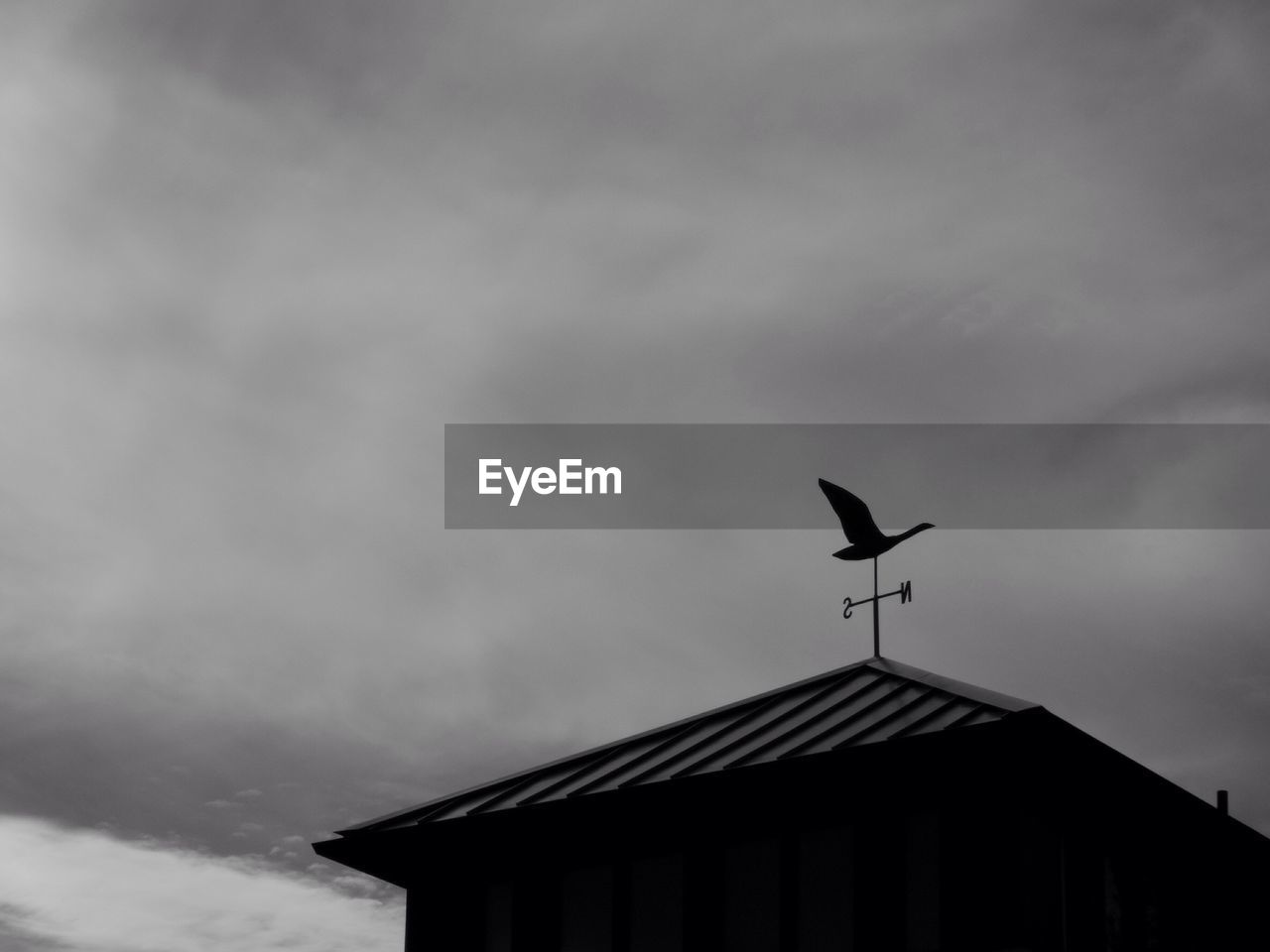 Low angle view of weather vane on roof against cloudy sky