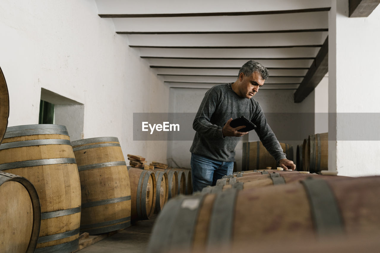 Winemaker holding digital tablet while examining wine barrels at cellar