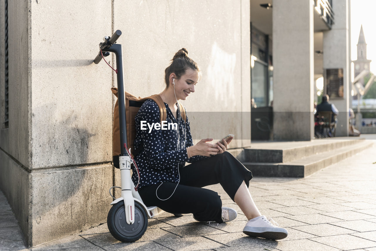 Smiling young woman with electric scooter, earphones and cell phone having a break in the city
