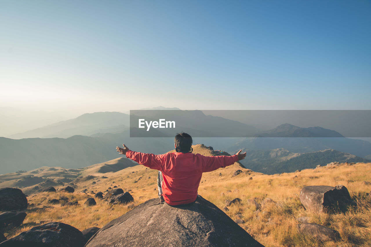 REAR VIEW OF MAN ON ROCK AGAINST MOUNTAIN