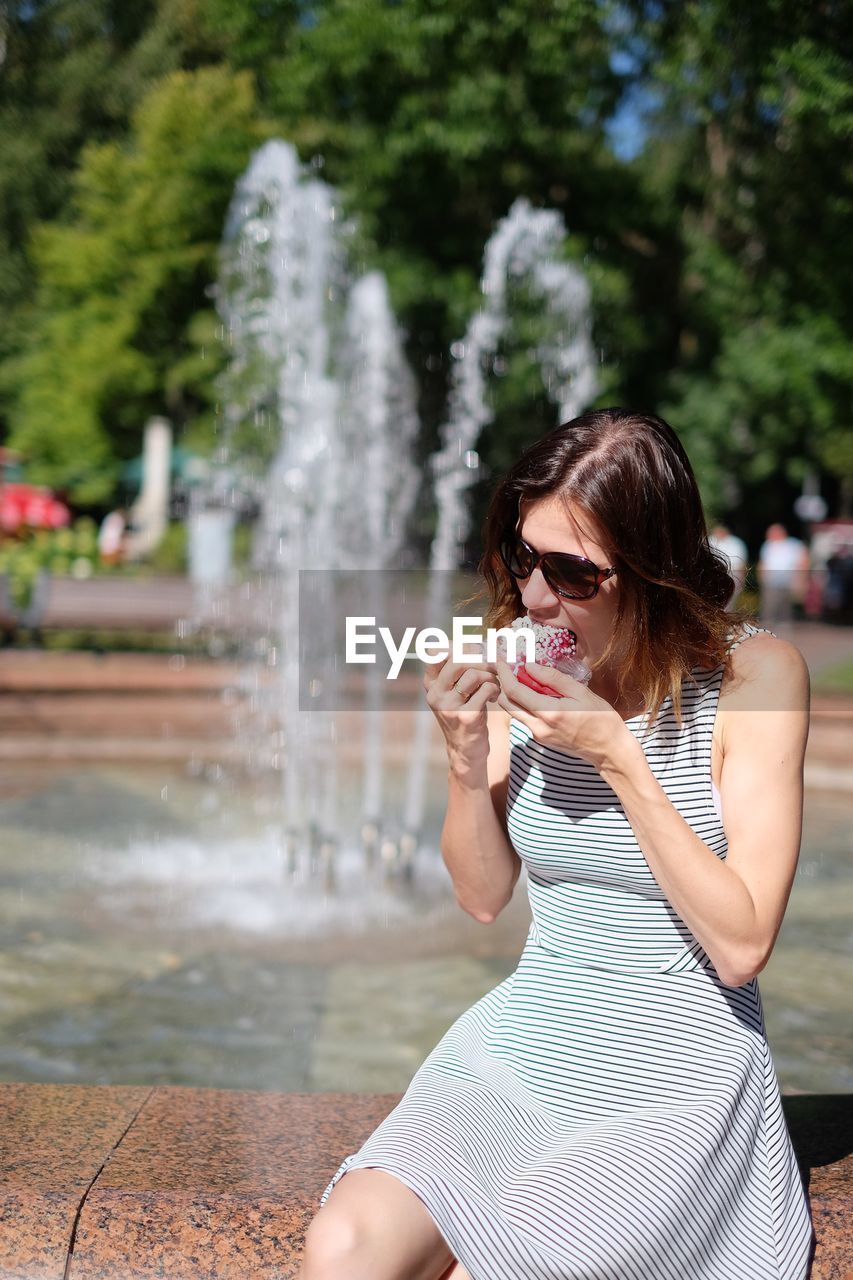 Mid adult woman eating slush while sitting on retaining wall against fountain