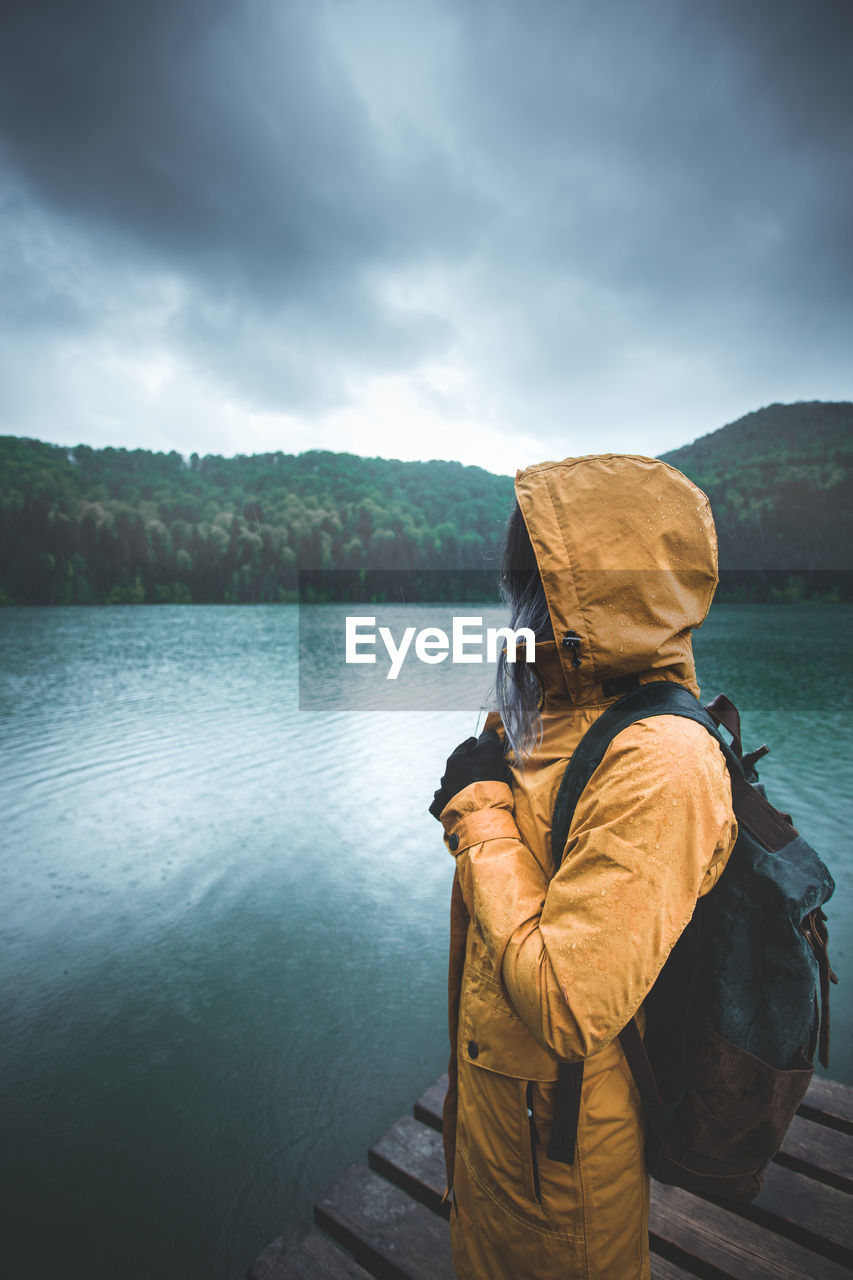 Young woman wearing raincoat while standing on pier by lake during rainy season