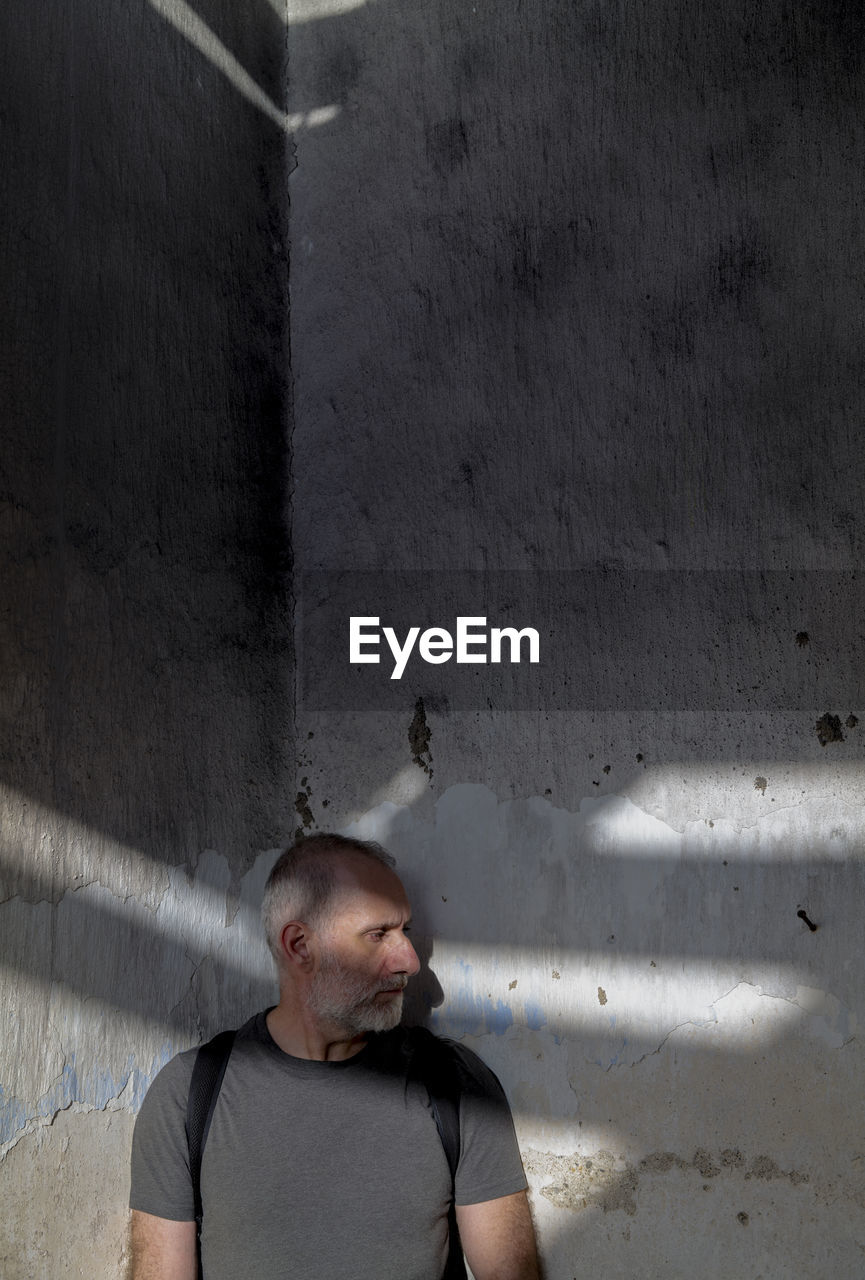 Portrait of adult man looking away while standing on old wall with sunlight and shadow