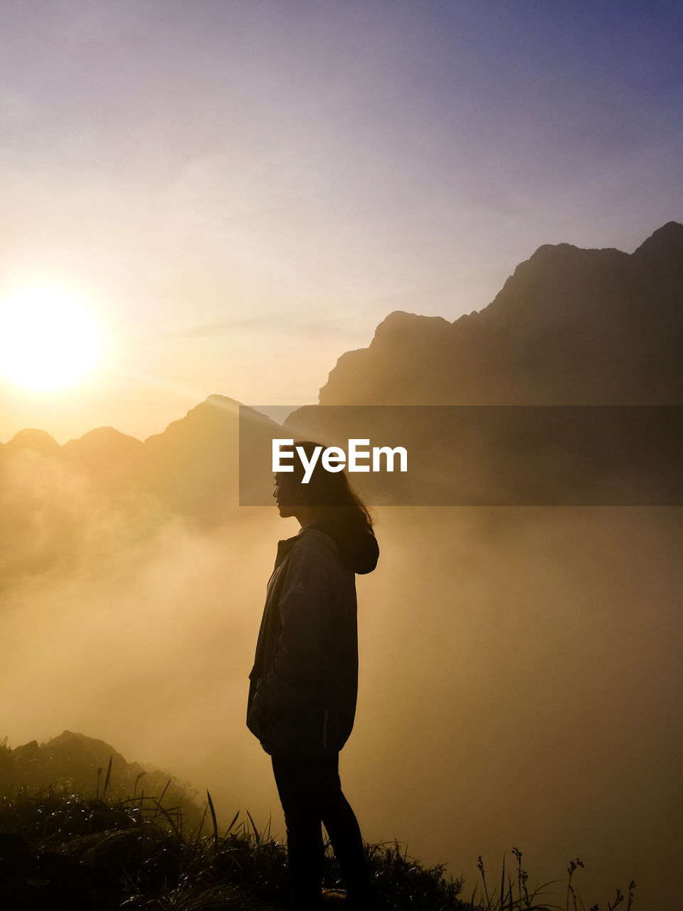 Side view of woman standing on mountain during foggy weather at sunrise
