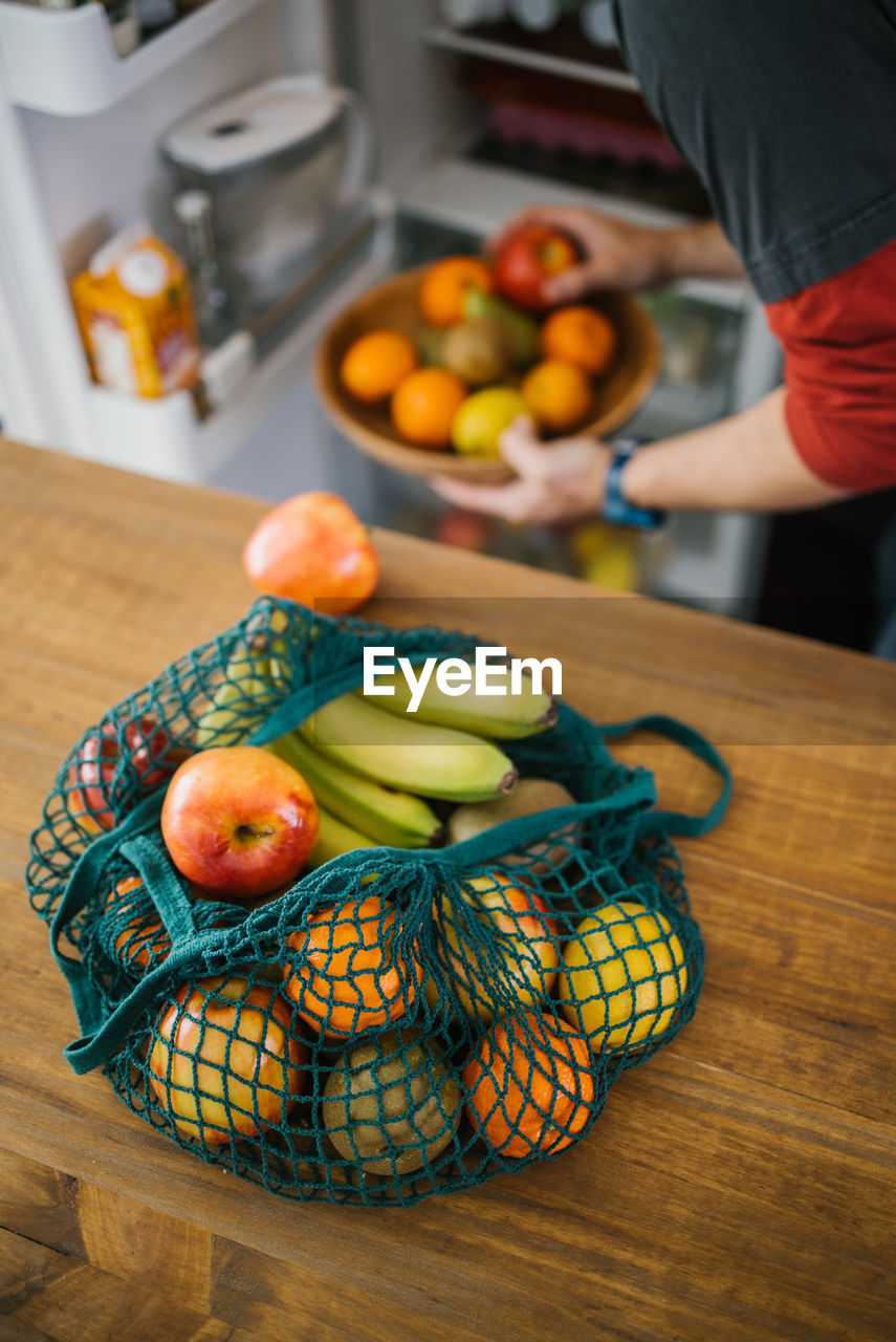 High angle of eco friendly cotton sack with ripe fruits placed on table in kitchen on background of crop person