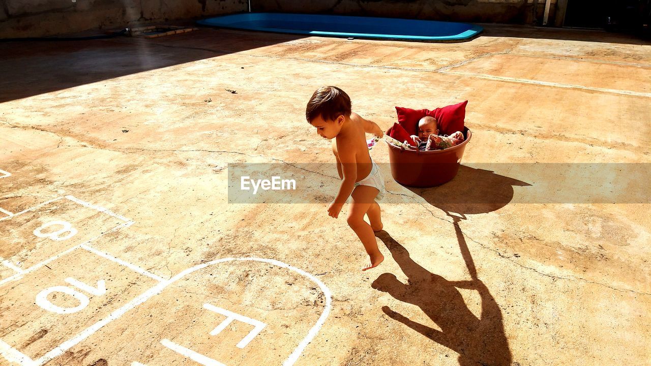 High angle view of shirtless baby boy pulling brother sleeping in bucket