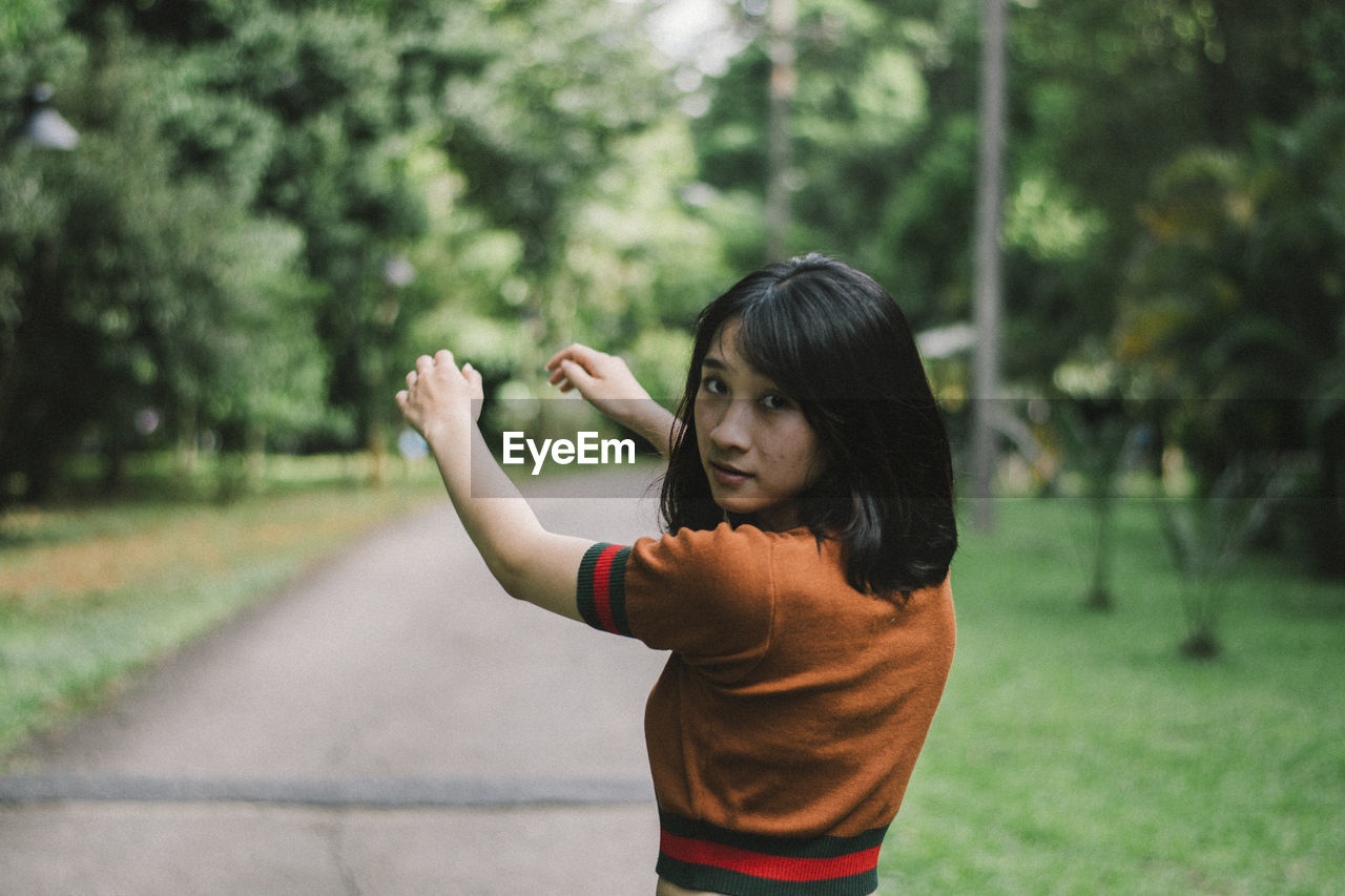 Portrait of young woman gesturing while standing on footpath in park