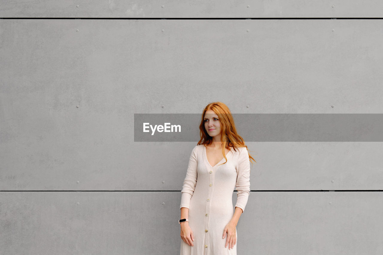Positive young female with long red hair wearing casual dress and smart bracelet looking away while standing against concrete wall