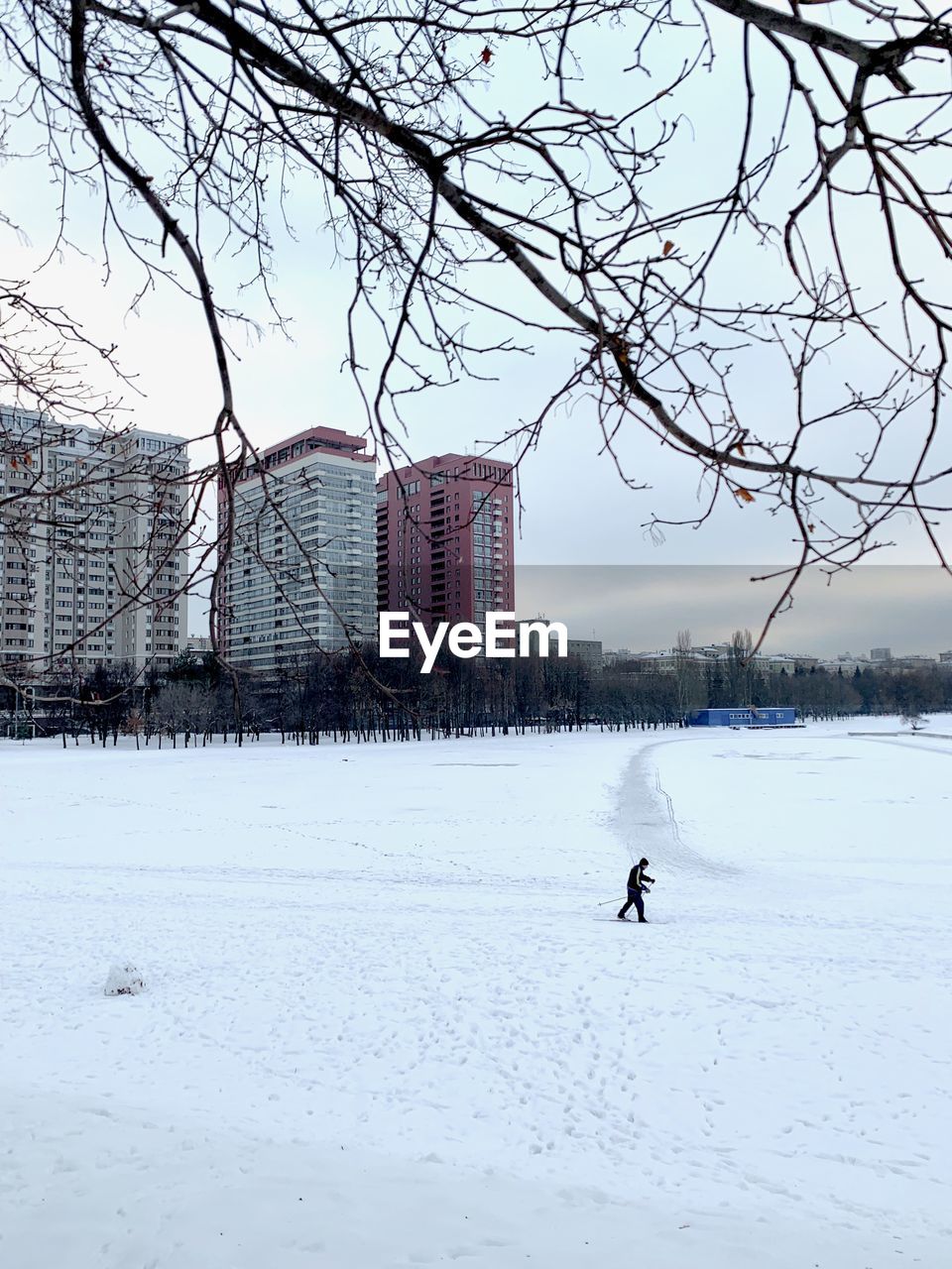 MAN WALKING ON SNOW COVERED FIELD BY BUILDINGS AGAINST SKY