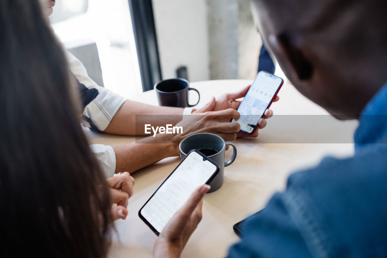 Group of cropped unrecognizable multiracial coworkers surfing cellphones while sitting at table with hot drinks during break in modern light cafe