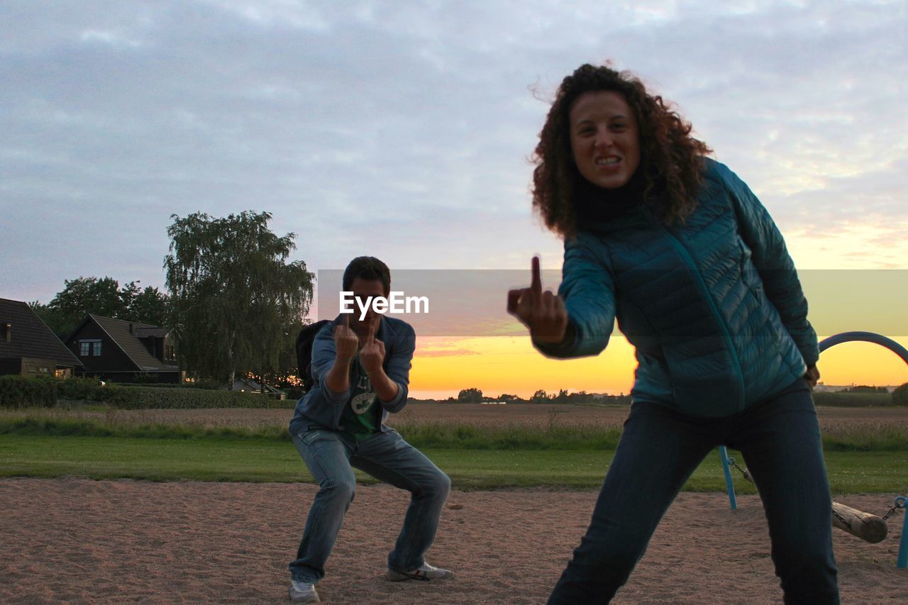 Portrait of friends showing middle finger at playground against sky during sunset