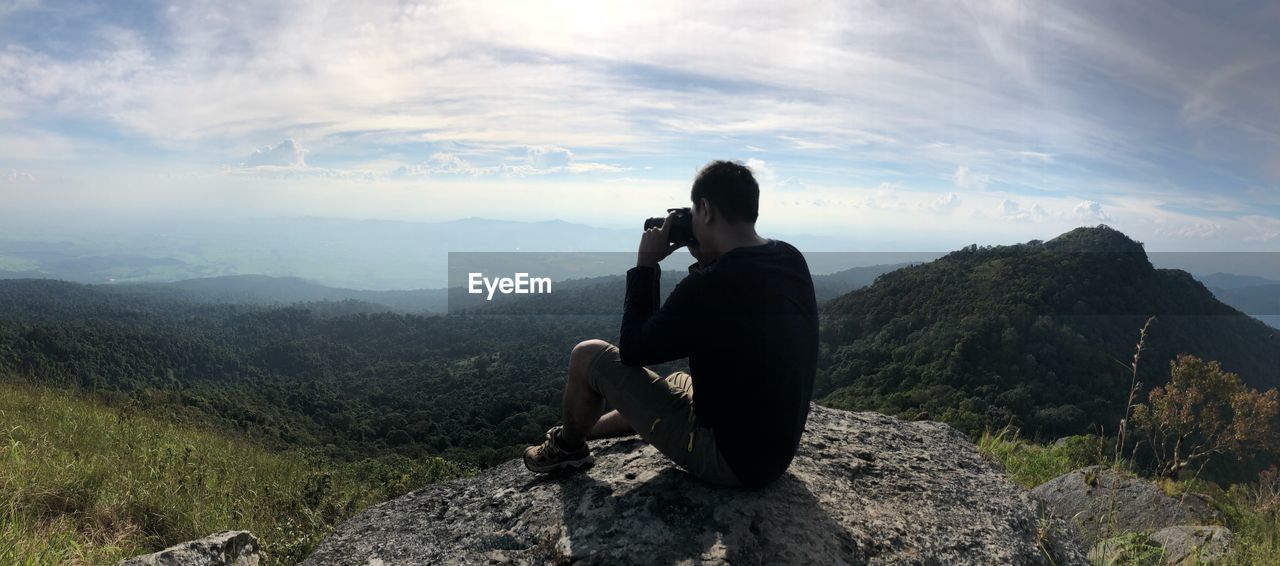 Man photographing while sitting on cliff against cloudy sky