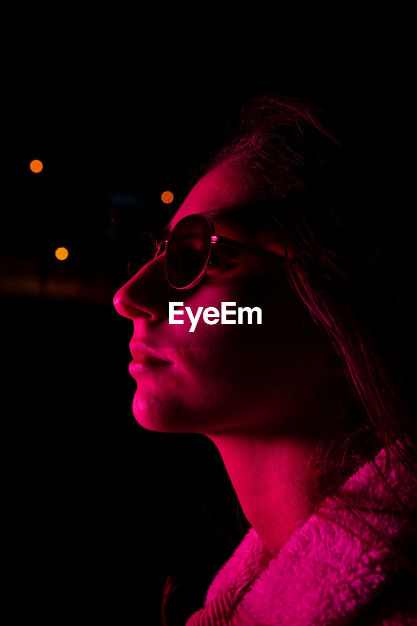 Crop young female in stylish sunglasses in dark studio with pink neon illumination and looking away with gentle smile