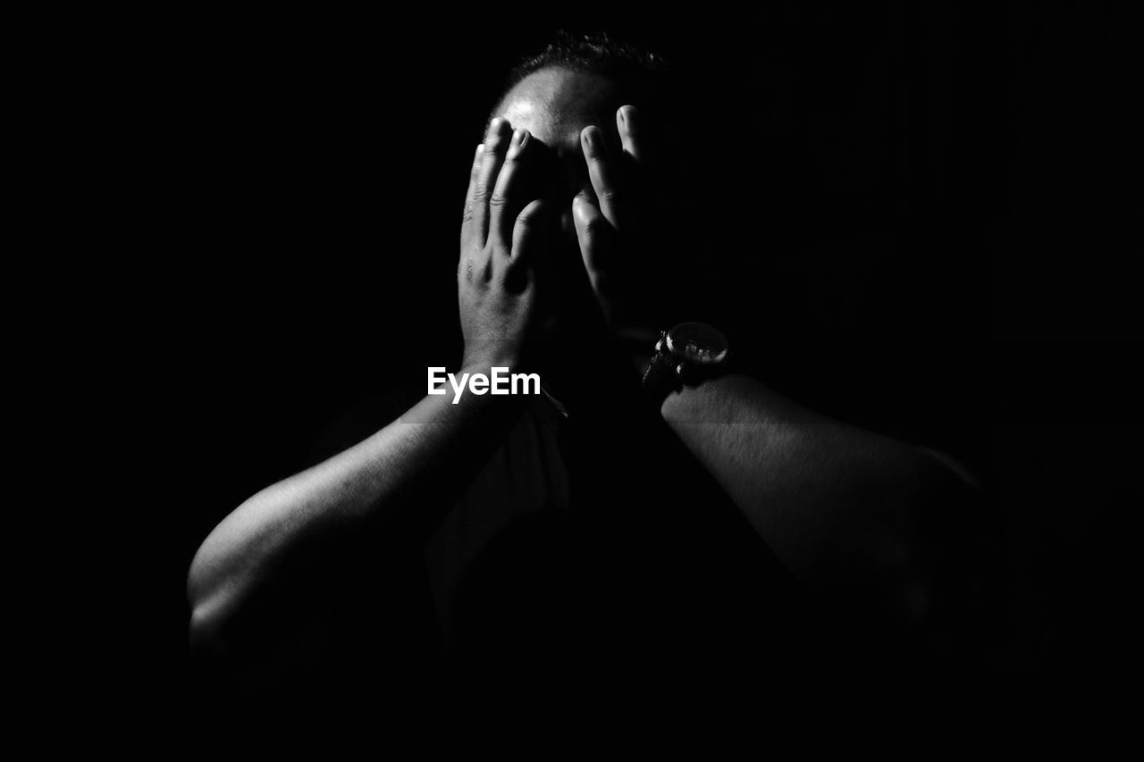 Close-up of man with hands covering face standing against black background