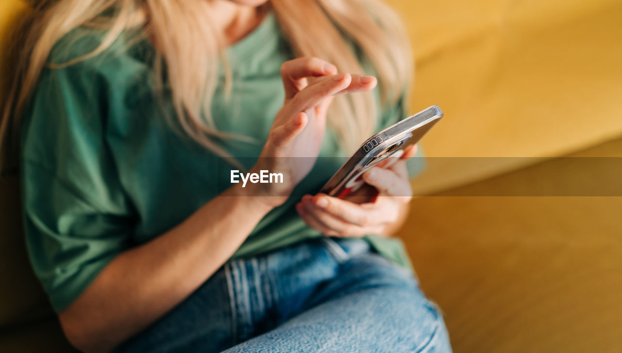 Unrecognizable woman browsing media on mobile phone while sitting on yellow sofa at home.