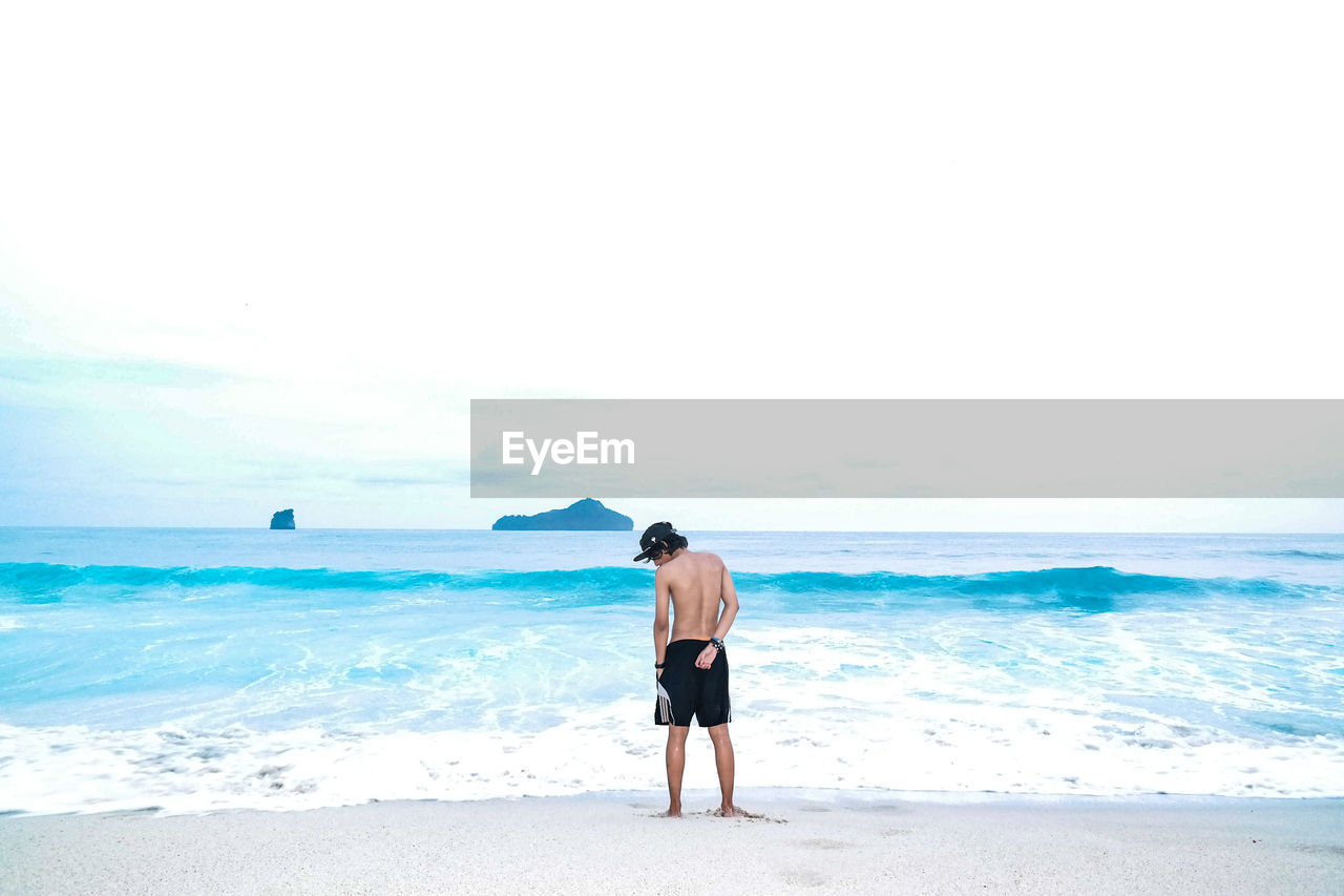 REAR VIEW OF SHIRTLESS MAN LOOKING AT SEA SHORE