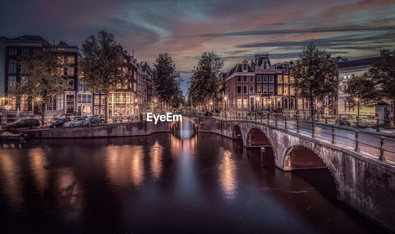 Bridge over river by buildings against sky at dusk