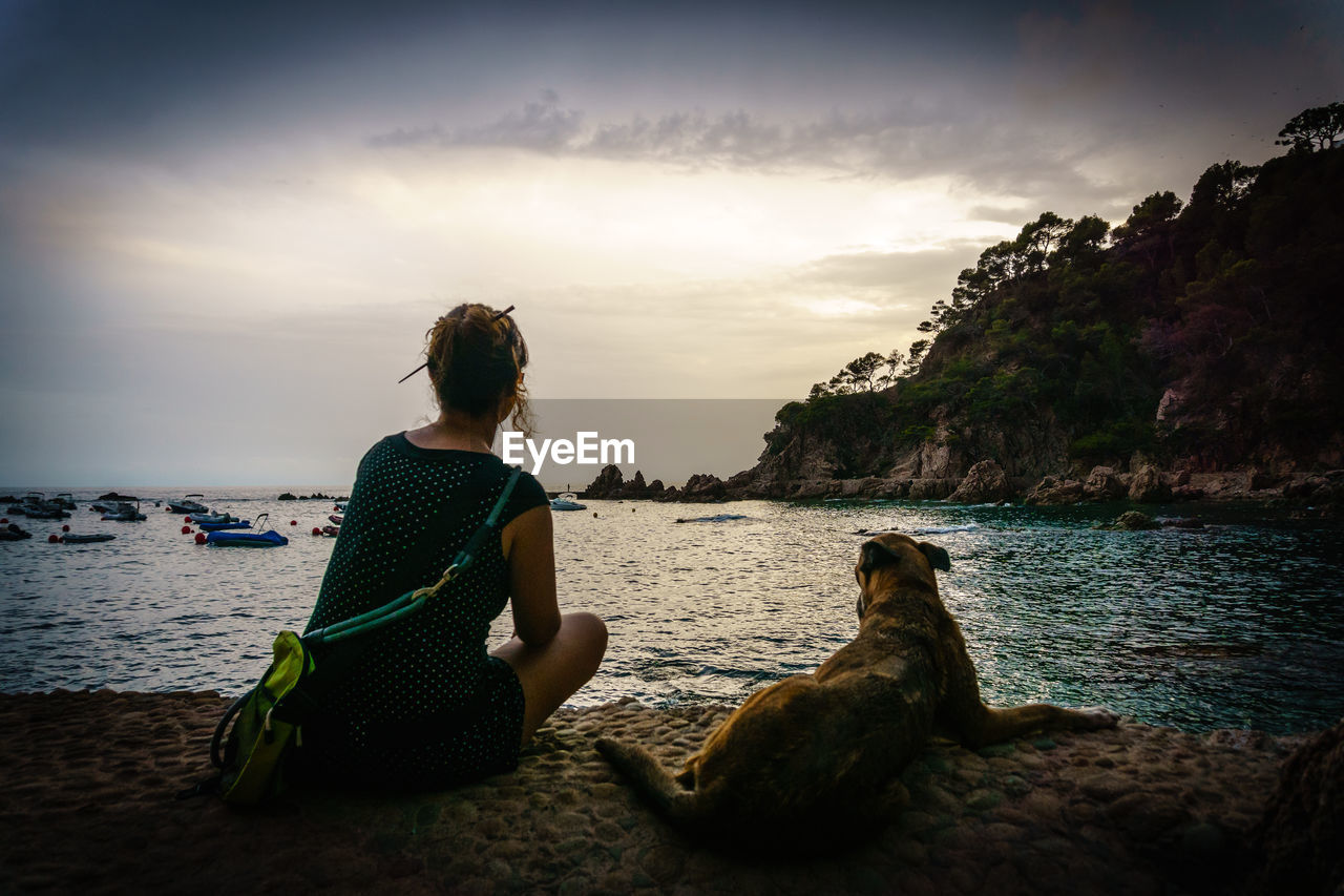 Rear view of woman with dog on beach against sky during sunset