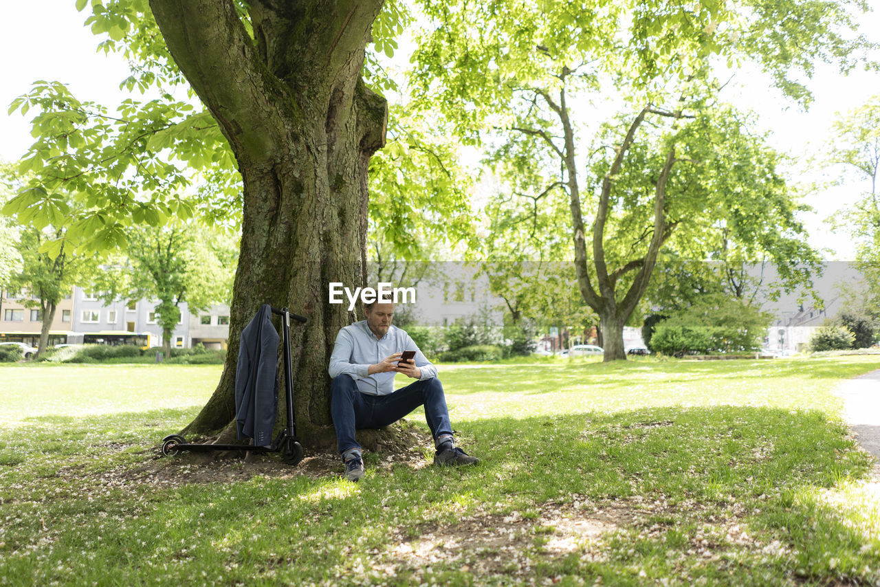 Businessman with e-scooter leaning against tree trunk at city park using smartphone