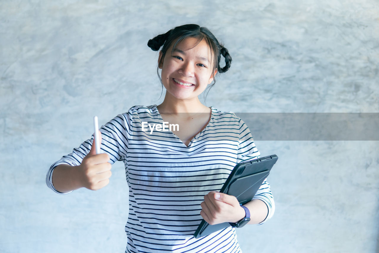 SMILING YOUNG WOMAN USING MOBILE PHONE AGAINST WALL