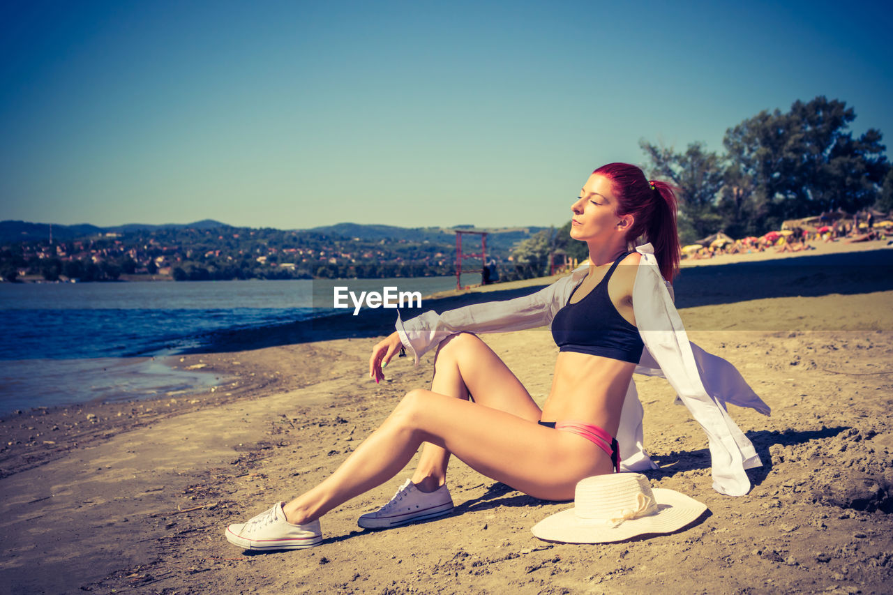 Redhead woman with eyes closed sunbathing at the beach by the river.
