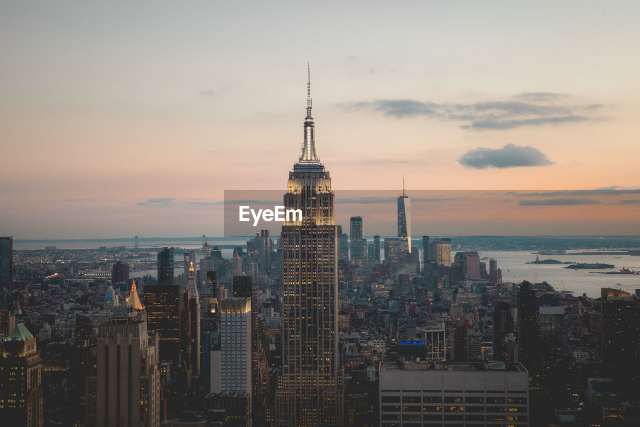 Empire state building amidst towers in city against sky during sunset