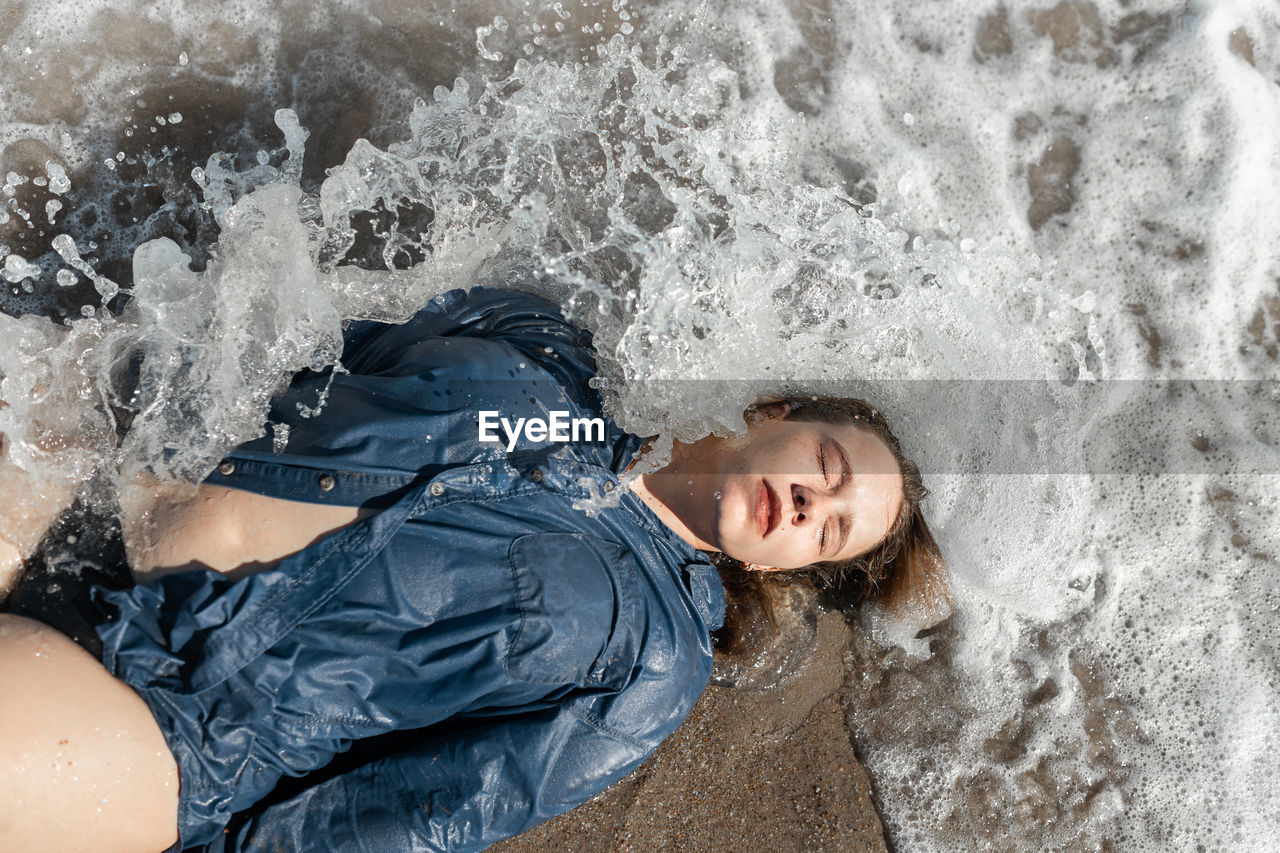 From above of calm female in wet shirt lying with closed eyes on sandy beach near waving sea on sunny summer day