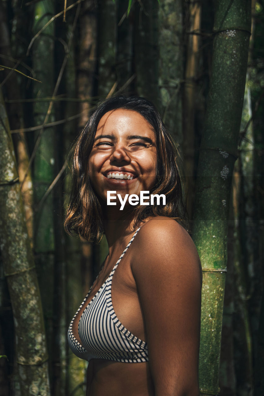 Side view of content young female traveler in swimwear with shadow on face looking away against bamboo twigs