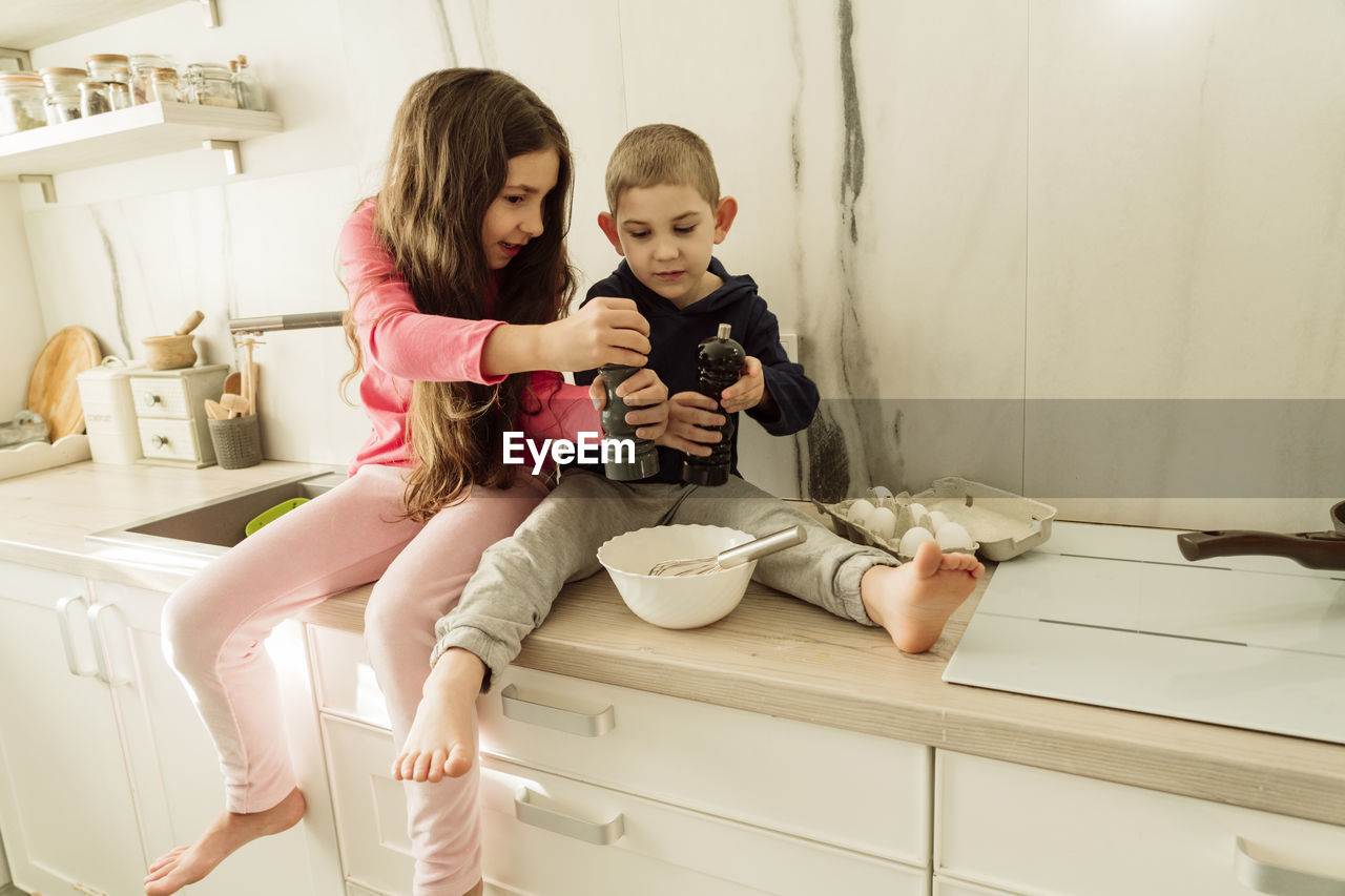 Girl with brother holding shakers over bowl sitting on kitchen counter at home