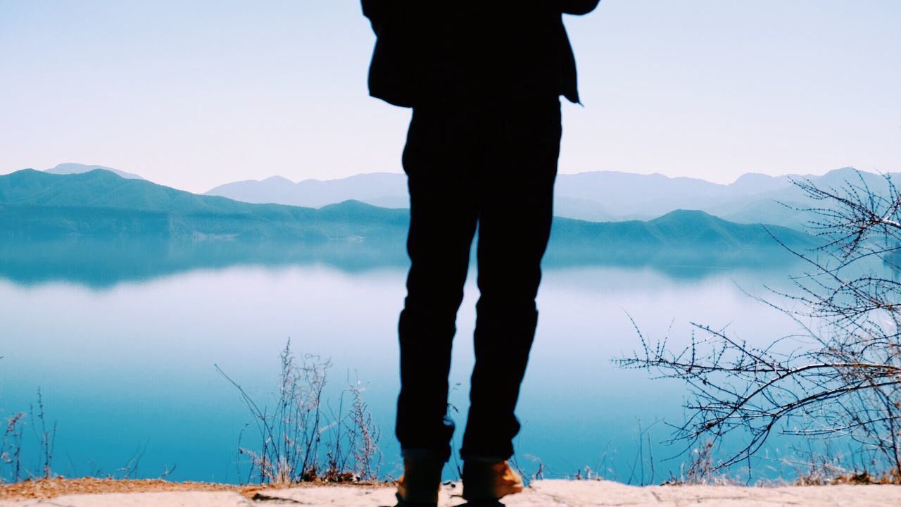 Low section of man standing at lakeshore against sky