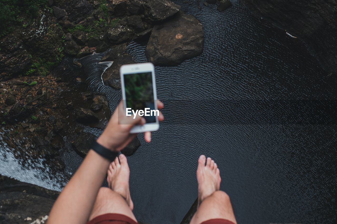 Low section of man photographing while sitting on cliff by waterfall in forest