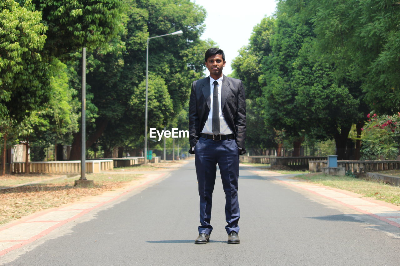 Full length portrait of young man wearing suit standing on road against trees