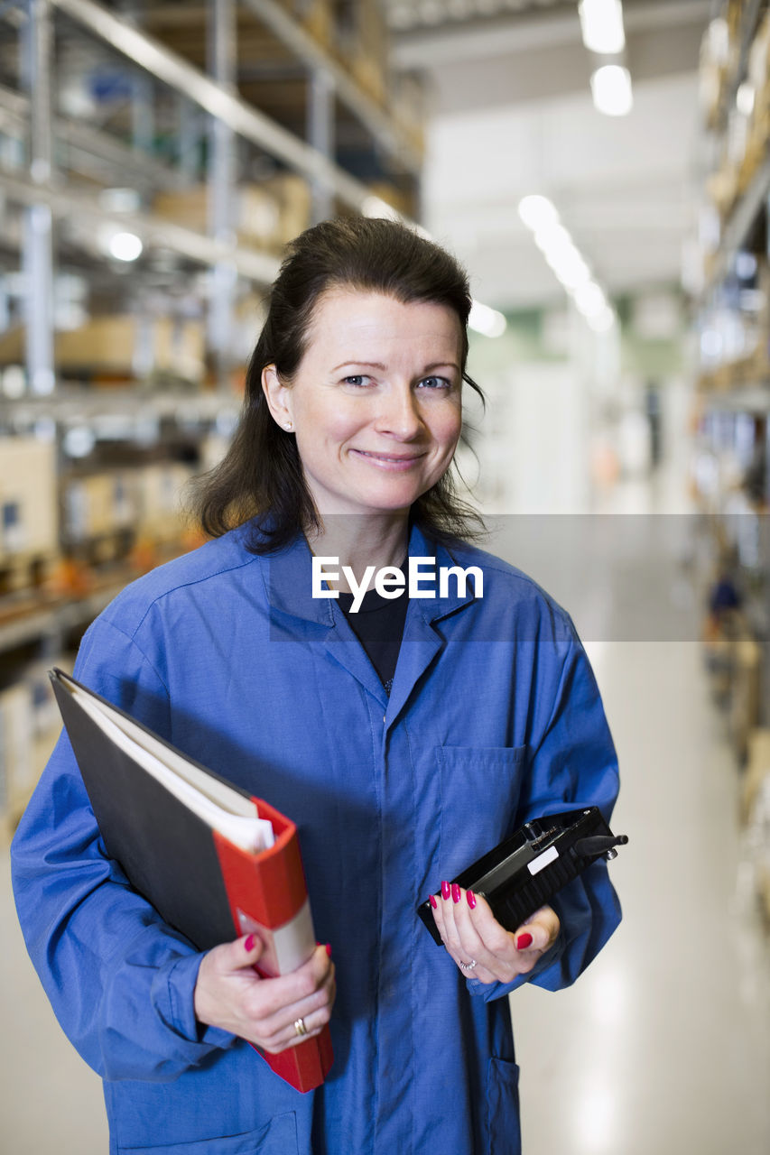 Portrait of happy female technician holding file and equipment while standing in industry