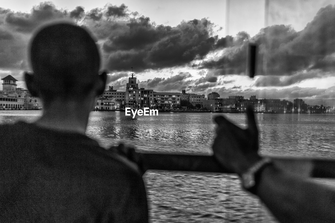 REAR VIEW OF MAN LOOKING AT CITYSCAPE BY RIVER
