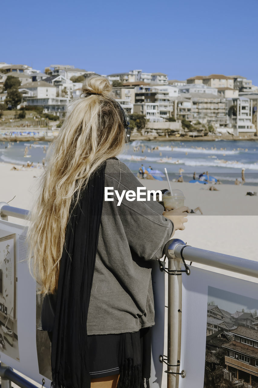 one person, winter, blond hair, architecture, rear view, women, built structure, building exterior, adult, long hair, hairstyle, nature, spring, day, standing, sky, waist up, city, clear sky, leisure activity, lifestyles, three quarter length, clothing, water, snow, coat, cold temperature, travel, person, building, travel destinations, outdoors, sunny, vacation, tourism, railing, holiday, casual clothing, trip