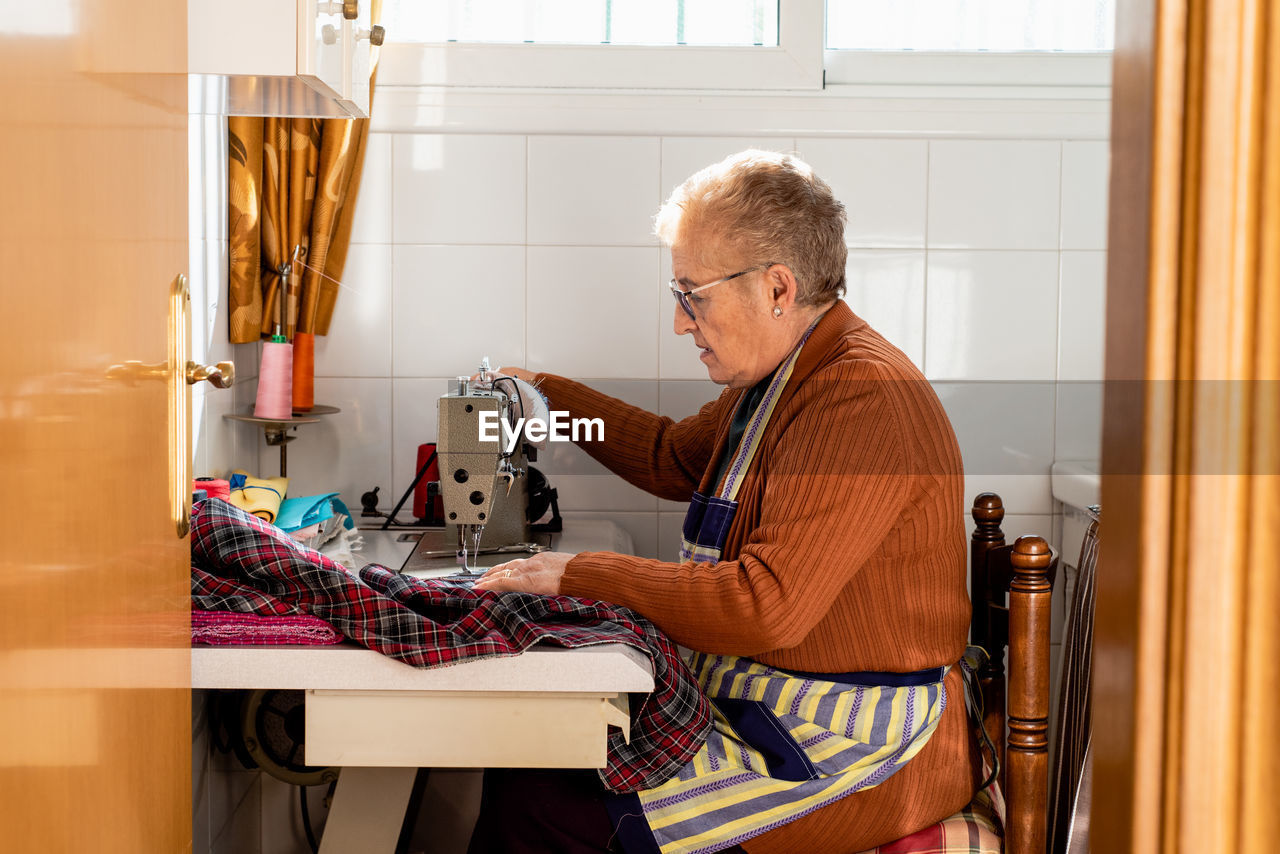 Side view of concentrated senior seamstress sitting at sewing machine and stitching piece of cloth while working in workshop