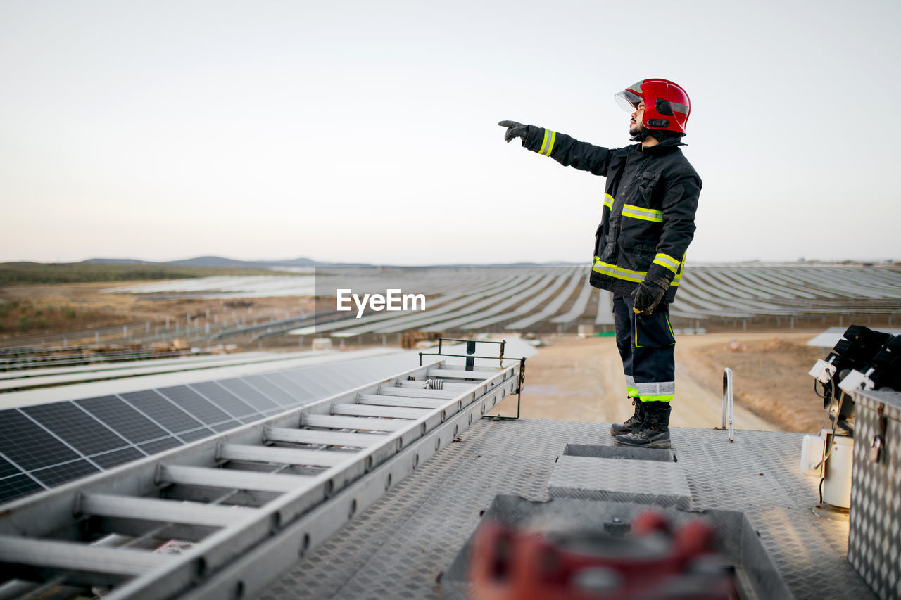 Full body side view of male firefighter wearing uniform and gloves standing on metal roof of fire engine near ladder with hand pointing forward at solar panels in rural farm