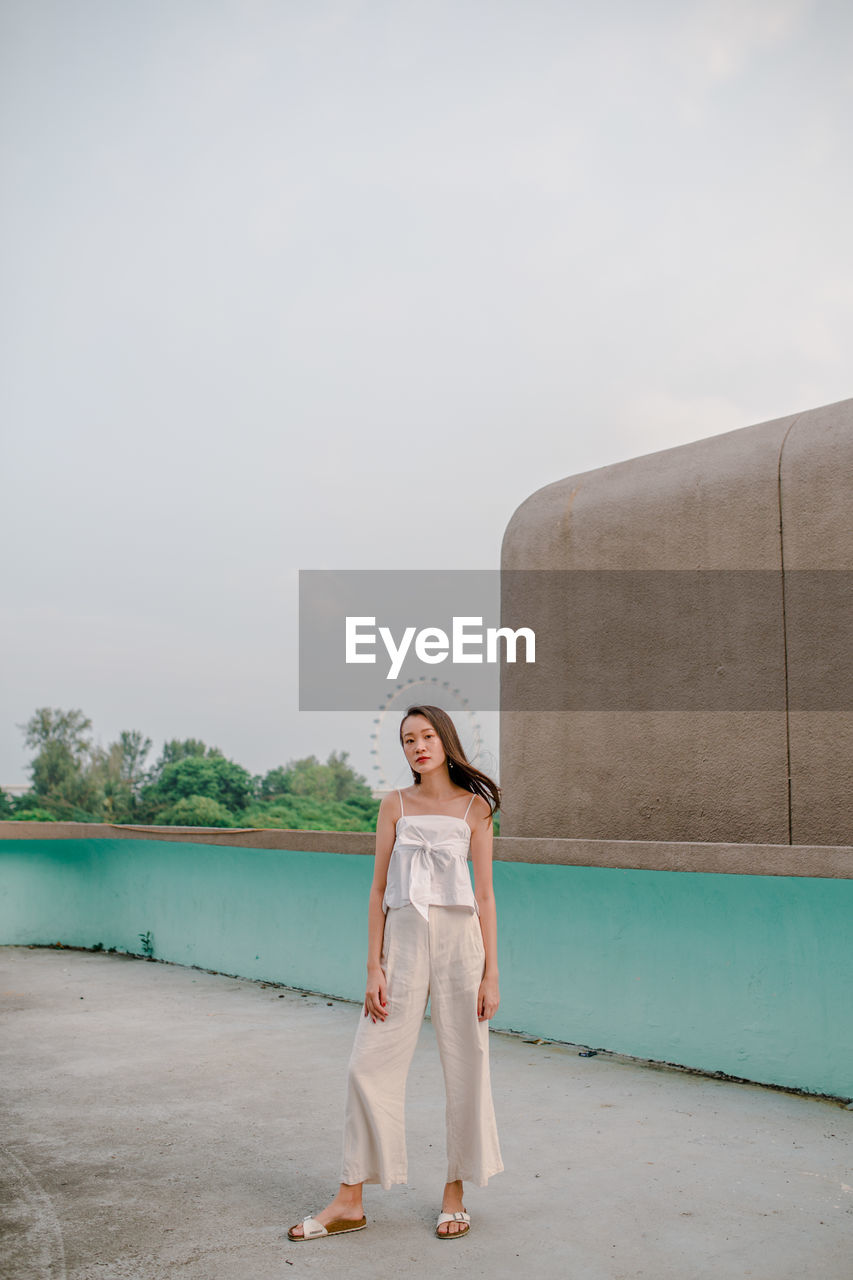 PORTRAIT OF YOUNG WOMAN STANDING AGAINST SKY