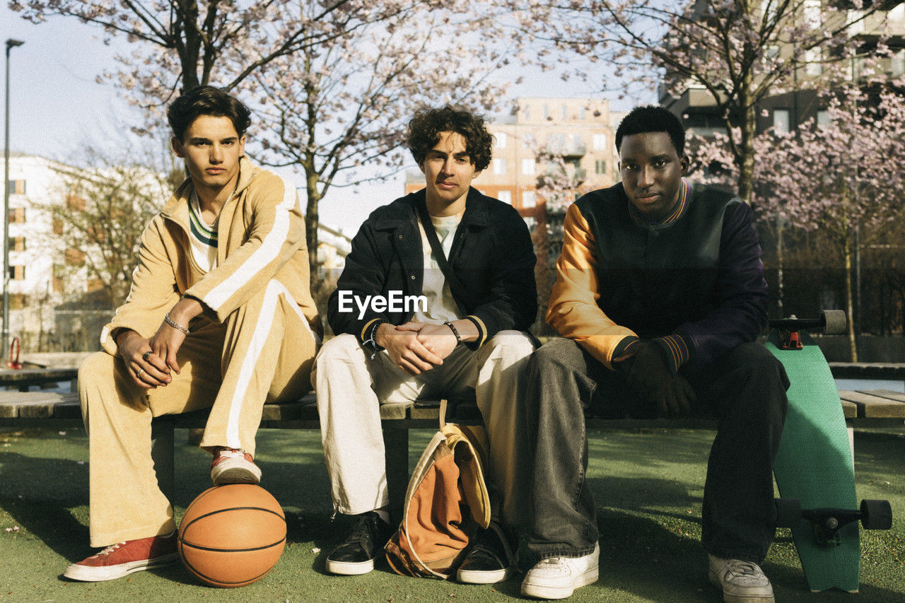 Portrait of young multiracial male friends sitting on bench during sunny day