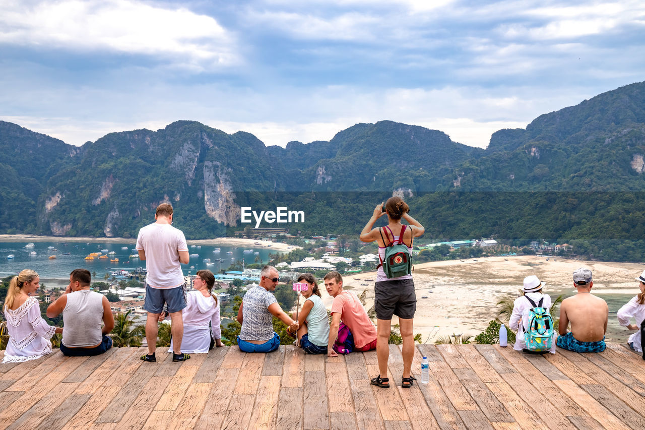 REAR VIEW OF PEOPLE SITTING ON SHORE AGAINST MOUNTAINS