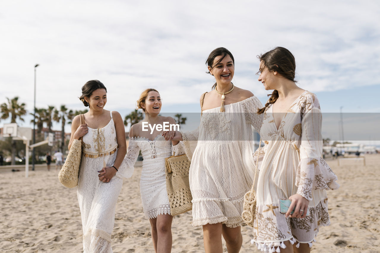 Young friends in white dresses smiling while walking at beach