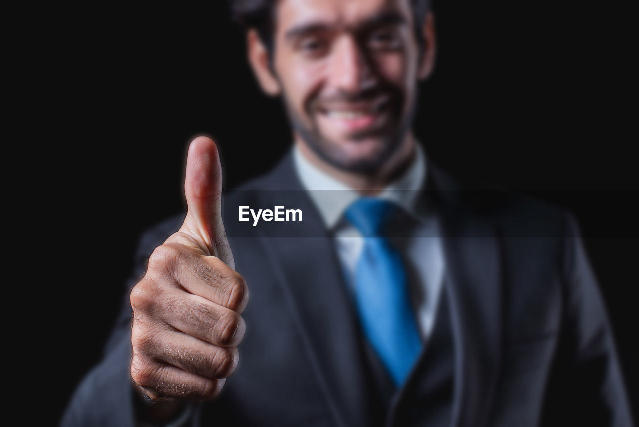 adult, men, one person, business, portrait, smiling, gesturing, businessman, formal wear, studio shot, emotion, thumbs up, happiness, looking at camera, success, hand sign, black background, front view, hand, person, cheerful, indoors, finger, sign language, professional occupation, pointing, corporate business, headshot, positive emotion, communication, menswear, white-collar worker, necktie, close-up, occupation, speech, showing, standing