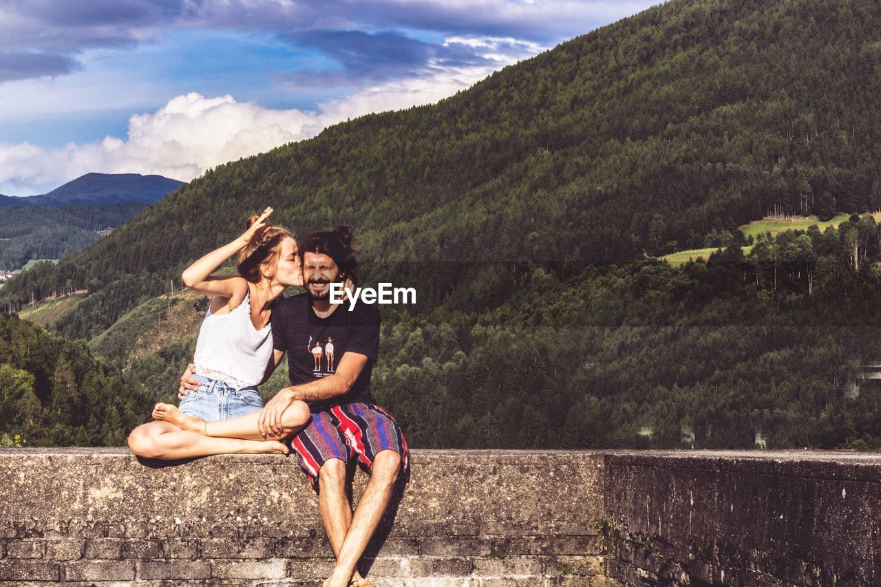 Woman kissing man while sitting on retaining wall