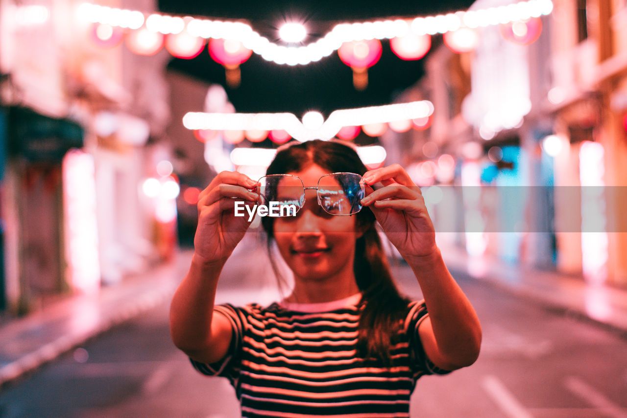 Young woman holding eyeglasses while standing on illuminated road at night