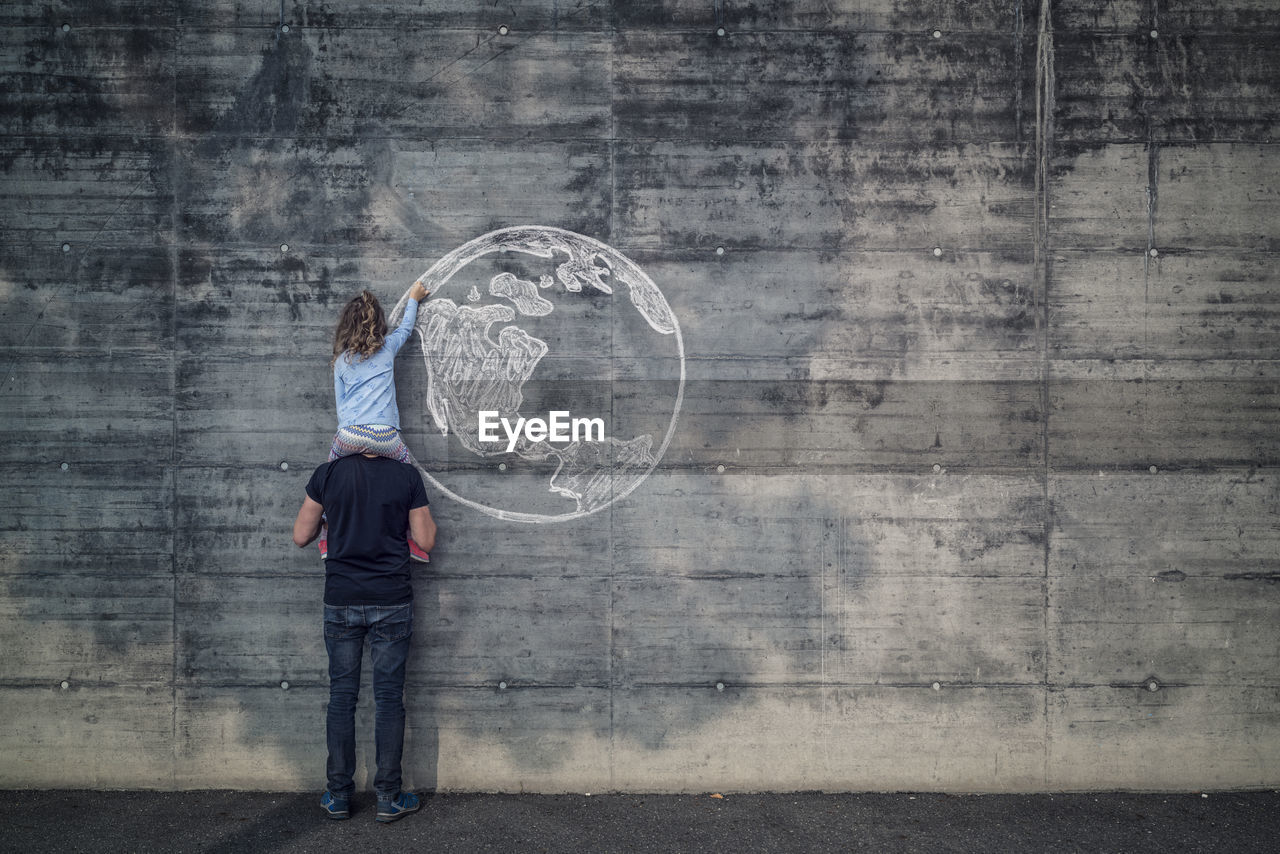 Austria, salzburg, father with daughter on his shoulders, the daughter draws with chalk the earth on a concrete wall