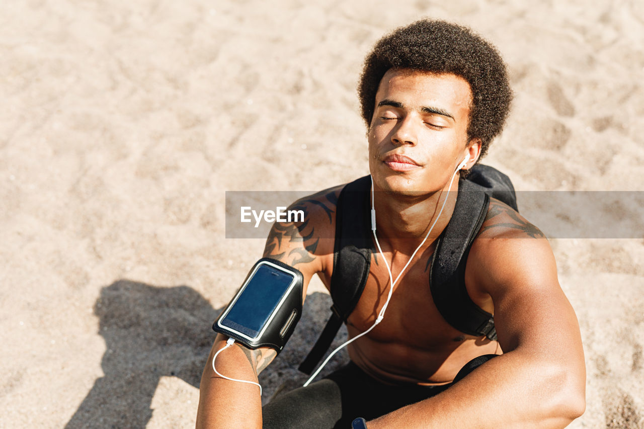 Shirtless male athlete listening to music through in-ear headphones while sitting at beach