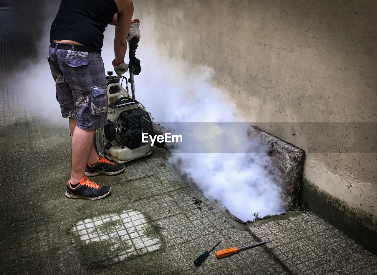 Low section of man standing by smoke emitting from machine against wall
