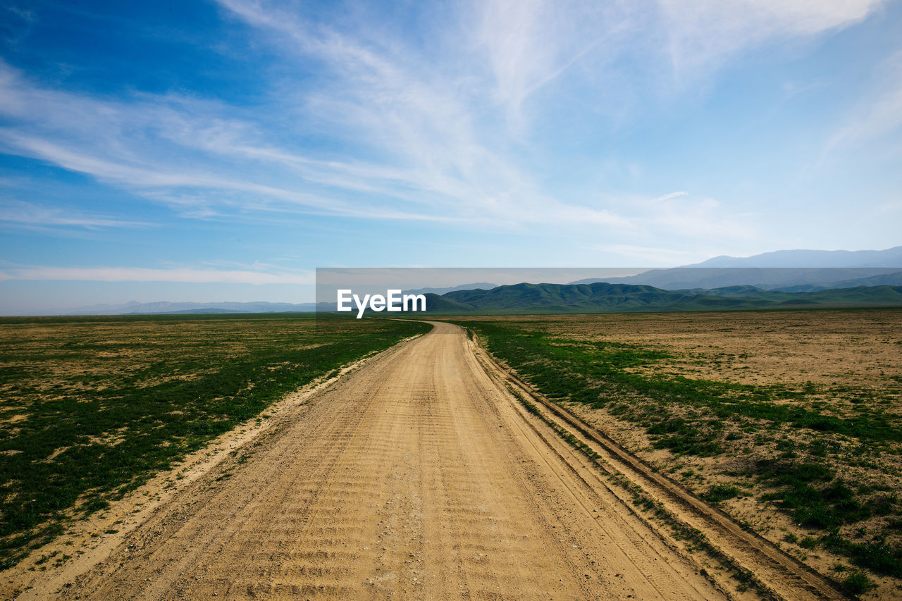 Empty dirt road amidst field against sky