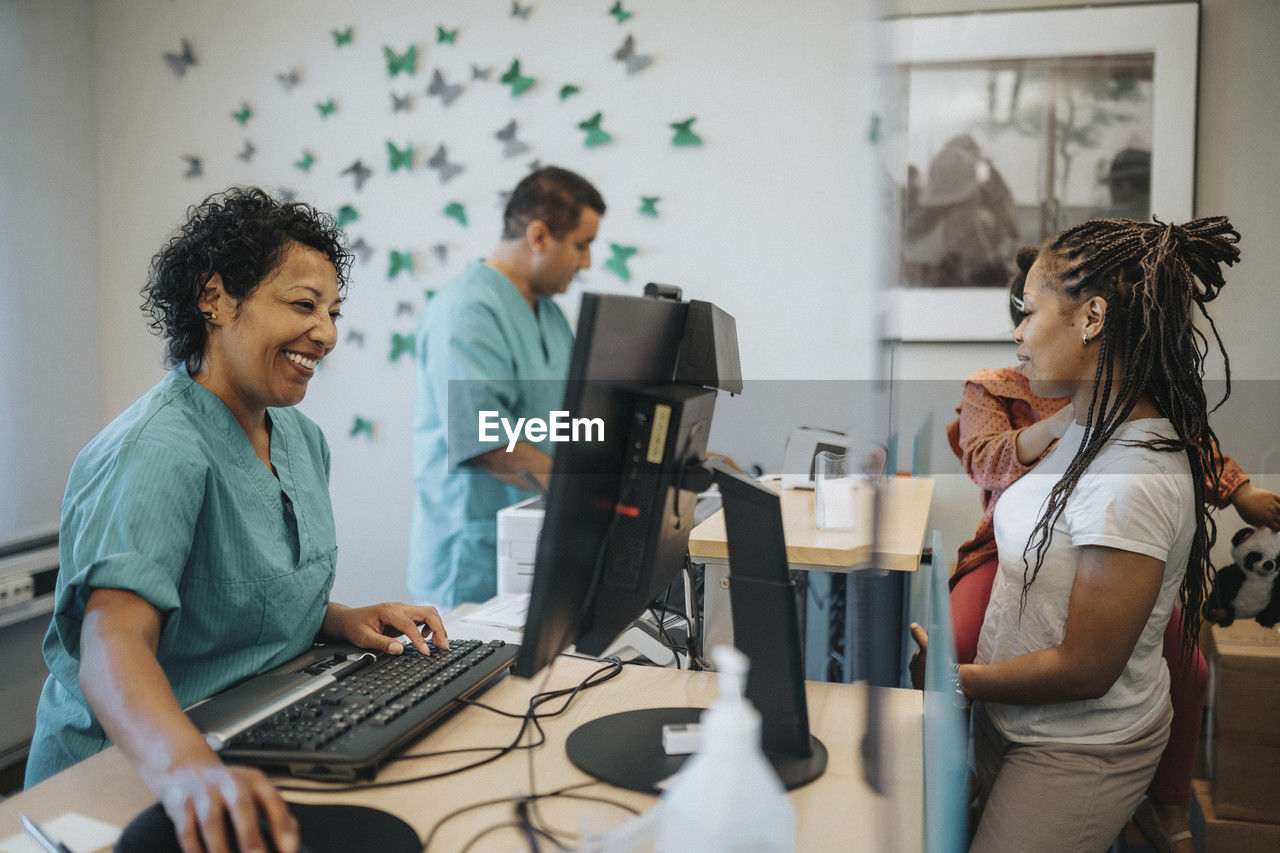 Happy female receptionist talking with mother carrying daughter at receptionist desk in hospital